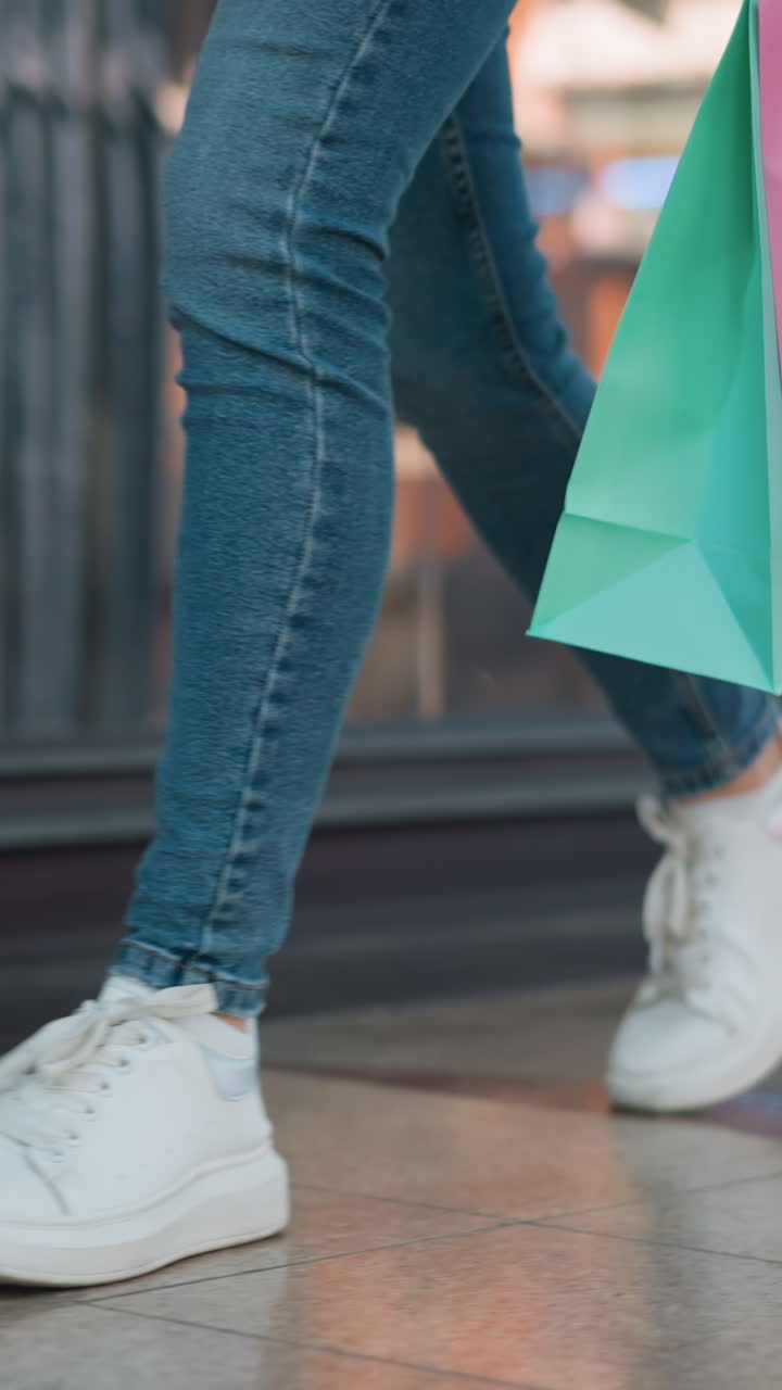 vista de la pierna de una persona con vaqueros y zapatillas de deporte de lona blanca sosteniendo bolsas de compras rosadas y menta mientras camina a través de un centro comercial más allá de una tienda de ropa, con fondo borroso de pantallas minoristas