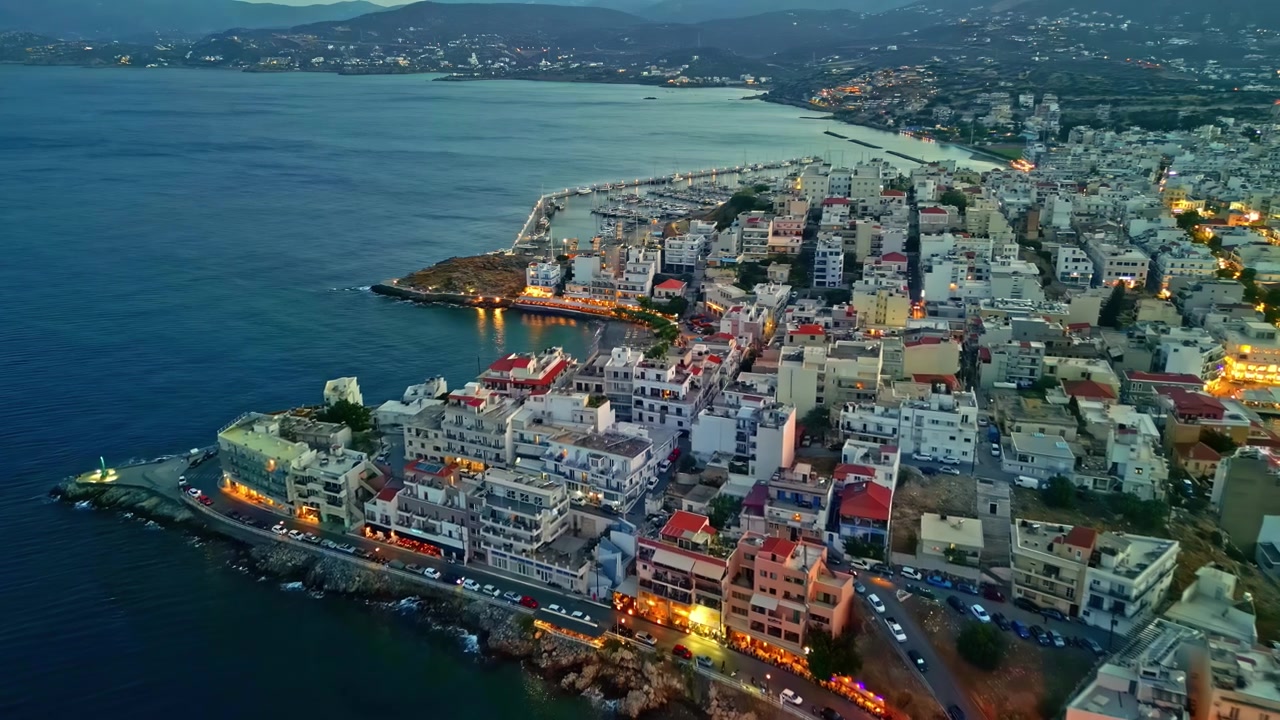 Luzes do crepúsculo delineiam a península de Agios Nikolaos enquanto um drone desliza por densos apartamentos à beira-mar e águas calmas do Mediterrâneo.