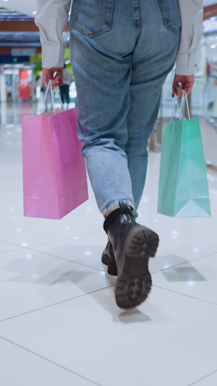 양손에 분홍색과 초록색 쇼핑백을 들고 현대적인 쇼핑몰을 통해 자신감 있게 걸어가는 세련된 여성의 뒷면, 그녀는 바쁜, 잘 조명 된 소매 공간을 통해 걸어갑니다.