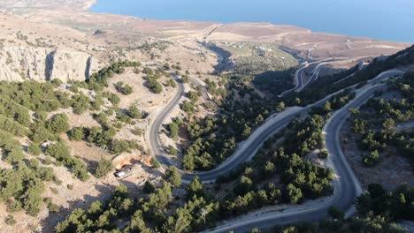 Drohnenansicht-In-Griechenland,-Die-An-Einem-Sonnigen-Tag-über-Einen-Braunen-Und-Grünen-Berg-Mit-Schlangenstraße-Und-Meer-Am-Horizont-Fliegt