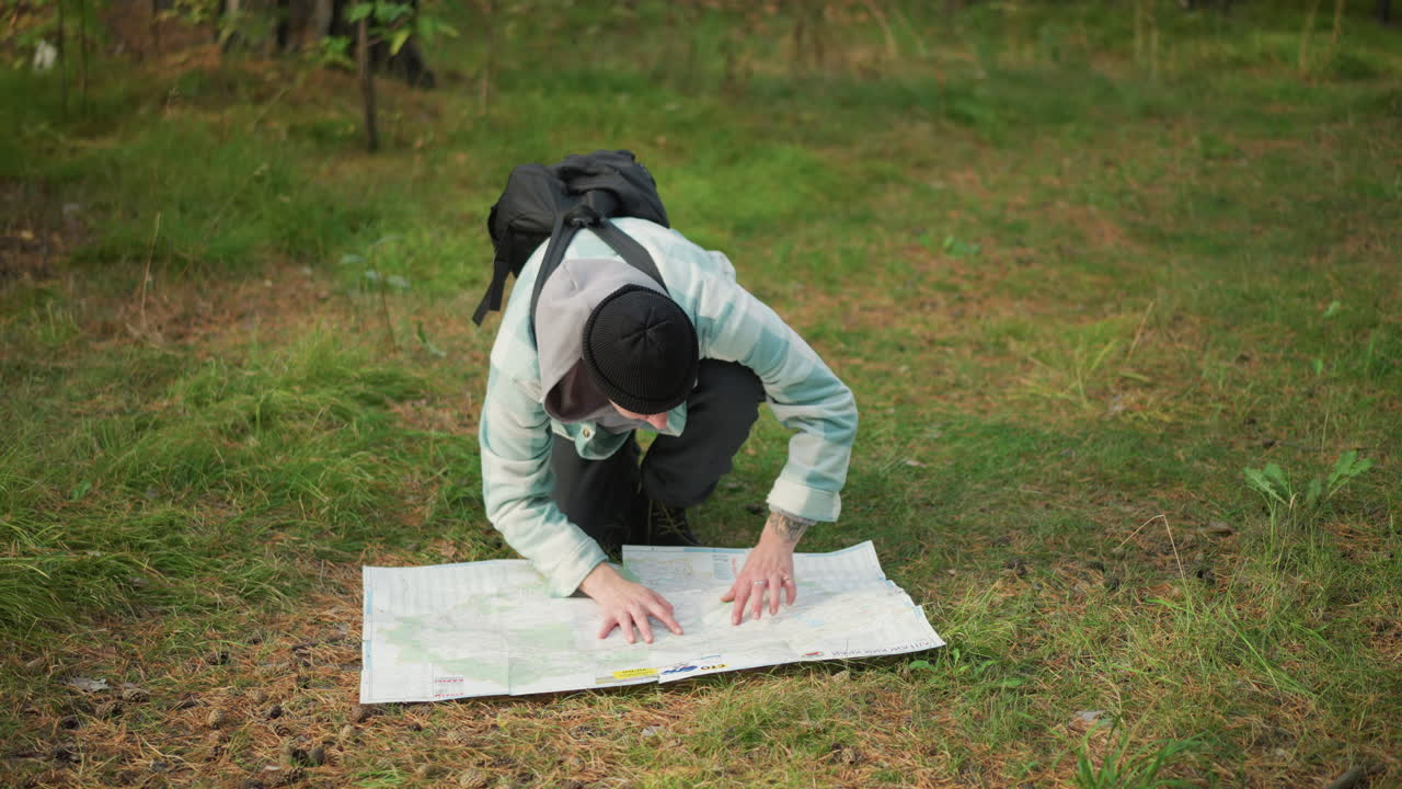 Caucasian hiker studying map on grass, kneeling in forest clearing with backpack and beanie, concentrating on route planning, folding and inspecting map, pointing to waypoints, compass nearby, calm