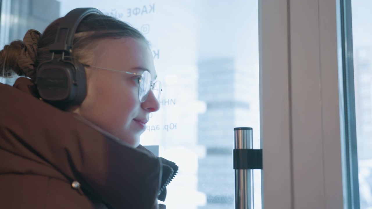Thoughtful Woman in Headphones Gazing Out Snowy Cafe Window