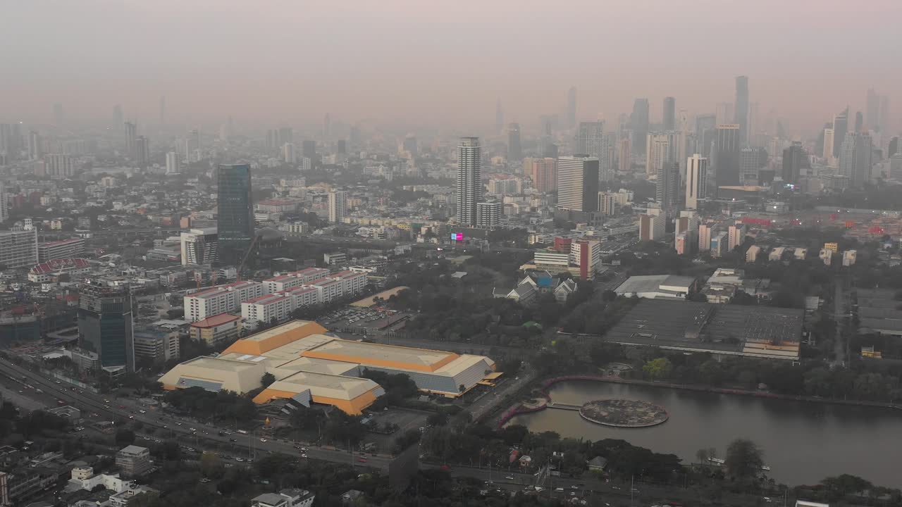 Vista de dron del Centro Nacional de Convenciones Reina Sirikit y el lago del Parque Benjakitti con los densos horizontes de Sukhumvit y Sathorn bajo un cielo brumoso, mostrando edificios de alta densidad urbana y contaminación atmosférica.