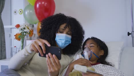 african american mother taking selfie with her daughter in oxygen mask lying on bed at hospital