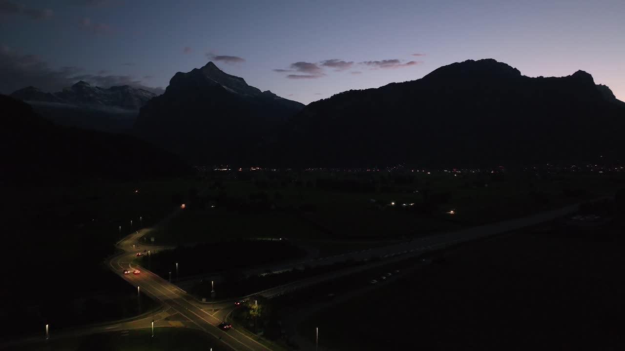 Aerial view of highway with illuminated vehicles passing against dark mountains near Amden, Weesen, Glarus, and Walensee, Switzerland, under clear sky.