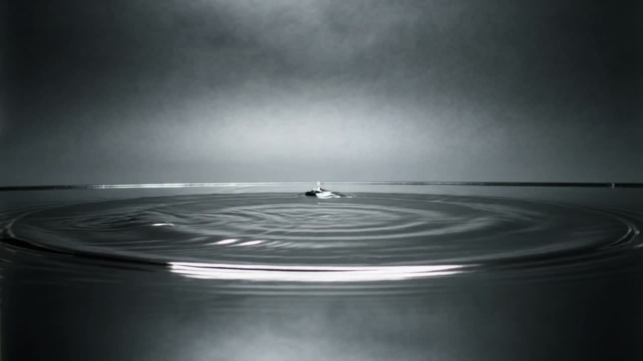 Impacting single drop creating rippling rings on shallow pool rim, showing rebound and lit backdrop