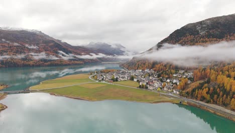las nubes bajas que envuelven a los vibrantes árboles de otoño en las laderas de las montañas que rodean silvaplana y su tranquilo lago crean una atmósfera serena en un día nublado.