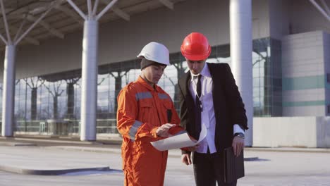 Junger-Architekt-Im-Schwarzen-Anzug-Mit-Krawatte-Untersucht-Das-Bauobjekt-Mit-Bauarbeiter-In-Orangefarbener-Uniform-Und-Helm.-Sie-Treffen-Sich-Am-Gebäude-Und-Schütteln-Sich-Die-Hände.