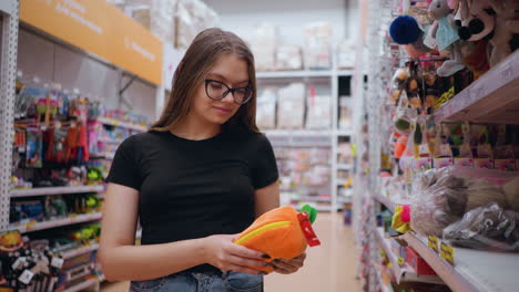 joven con camiseta negra y vaqueros recogiendo una bolsa de peluche naranja brillante de la estantería de una tienda en un centro comercial bien iluminado, examina el artículo mientras navega por la sección de juguetes llena de accesorios coloridos