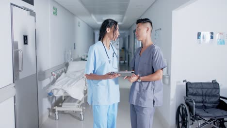 Diverse-doctor-and-nurse-using-tablet-and-talking-in-corridor-at-hospital,-in-slow-motion