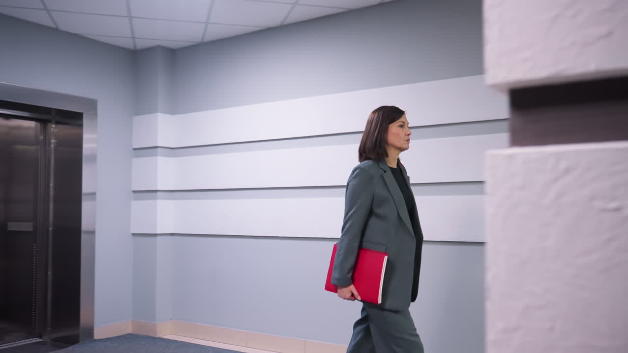 Businesswoman walking confidently in modern office hallway near elevator, carrying folder, dressed in formal business suit. Corporate workspace, professional environment, career-focused atmosphere