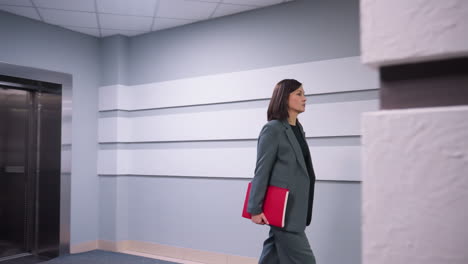 businesswoman walking confidently in modern office hallway near elevator, carrying folder, dressed in formal business suit. corporate workspace, professional environment, career-focused atmosphere