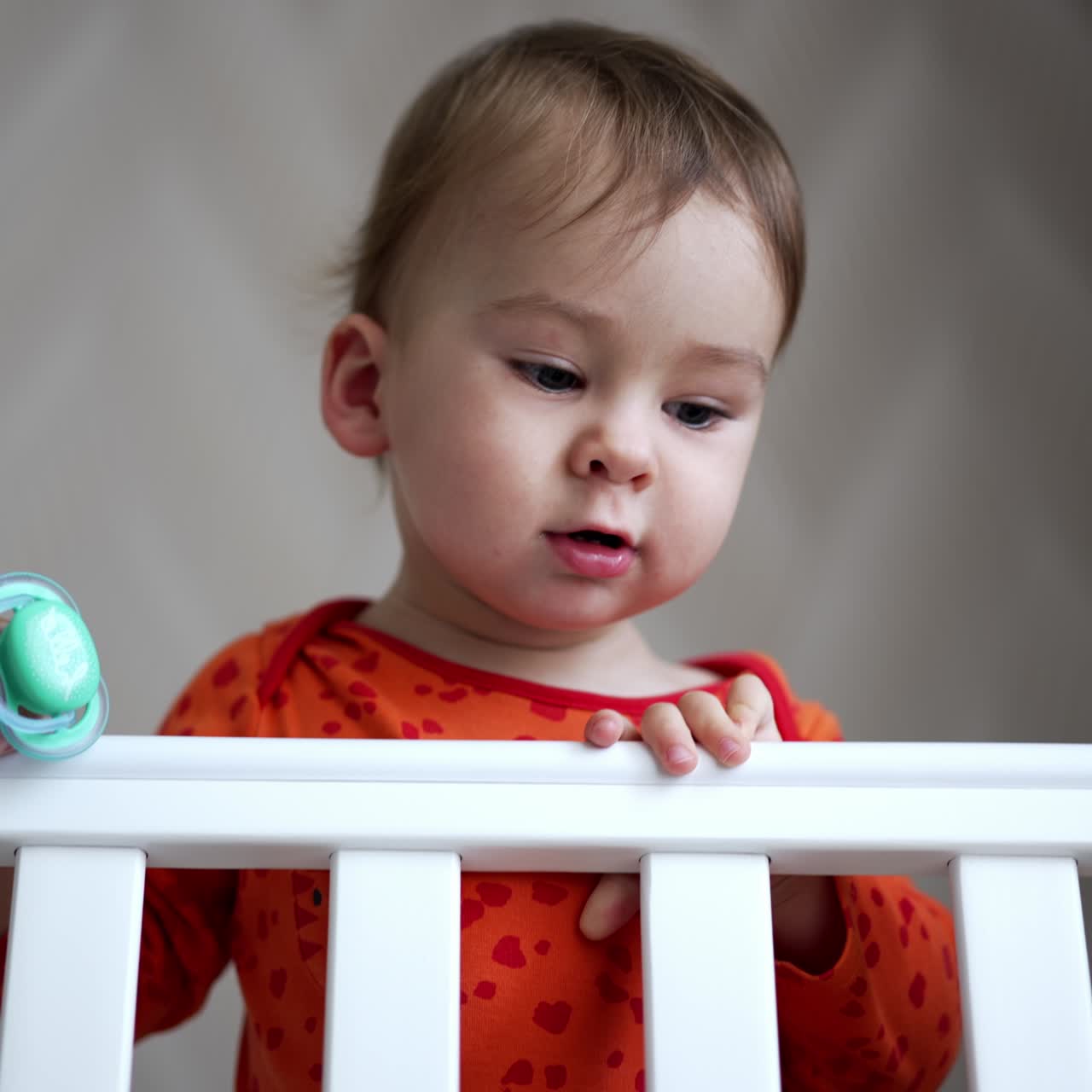 Lovely baby with cute plump cheeks standing in the cot. Adorable little boy holding a pacifier in hand. Low angle view