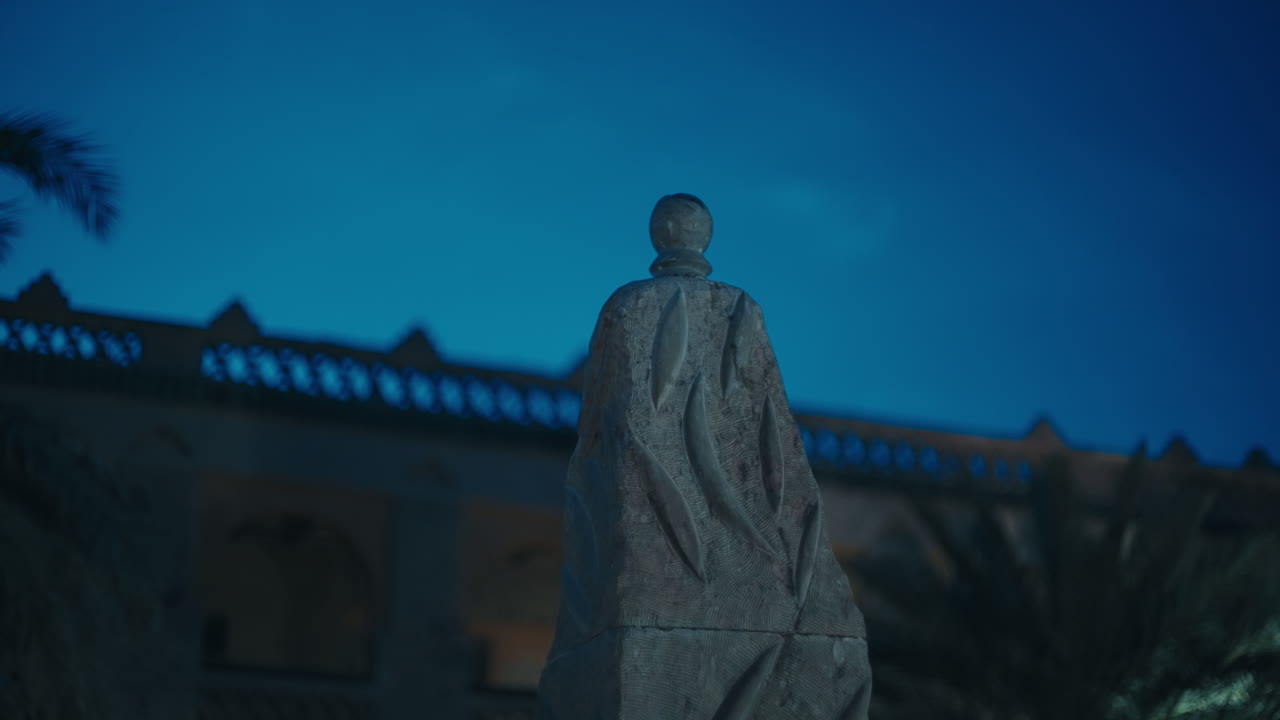Steinmonument eller skulptur som står utenfor en opplyst kasbah-vegg under en dypblå nattehimmel i Marokko.