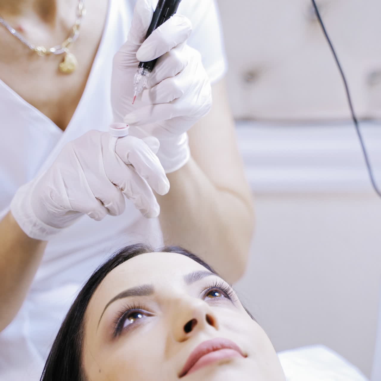 Skilled beautician fiiling up tattoer machine with red ink to make permanent make-up. Cosmetologist preparing for procedure of permanent make-up on lips for a young female in a beauty salon.