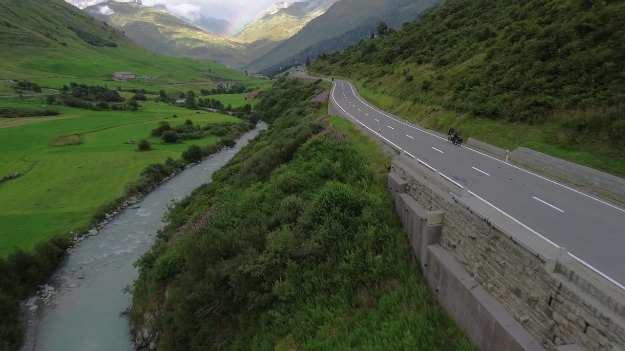 Cyclists on a road in Swiss Alps with cars and rainbow at the sky shot from a drone