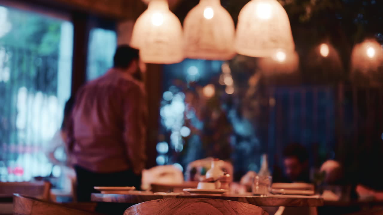 Una foto sfocata che mostra una persona in piedi in un ristorante con illuminazione calda, con tavoli e decorazioni in primo piano