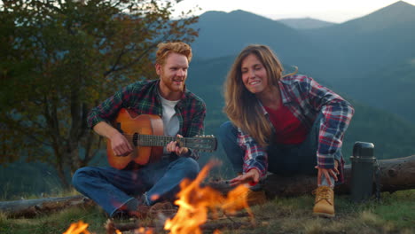 una pareja juguetona pasa las vacaciones sentada en una hoguera. los amantes tocan música en las montañas