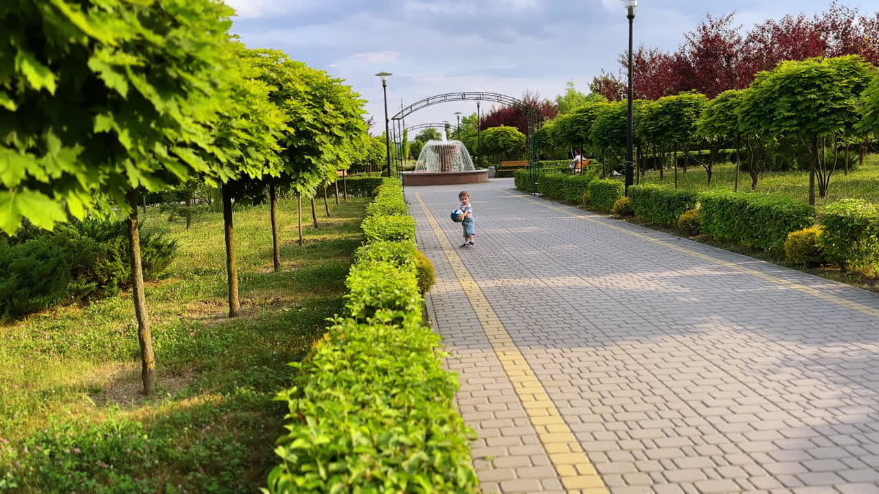 Little kid in jeans romper distancing from camera. Toddler runs with a ball in the green park in summer.