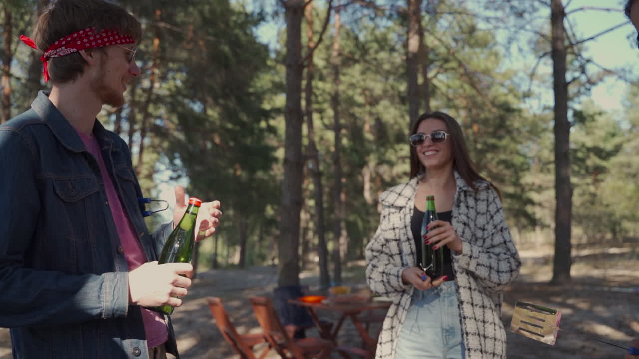 젊은 여성은 시골에서 바베큐를 하는 두 남자 친구에게 맥주 병을 줍니다. 사람들은 자연에서 피크닉을 즐기고 있습니다.