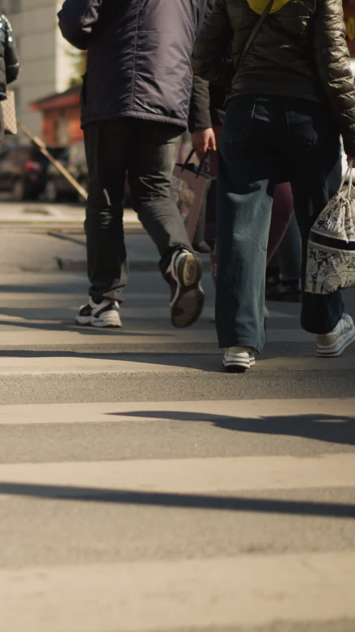 Trafficato attraversamento pedonale urbano con diversi pedoni, primo piano dal busto in giù di gambe e piedi che camminano sulle strisce zebrate, luce solare che proietta lunghe ombre sull'asfalto, zaini e giacche, scarpe da ginnastica