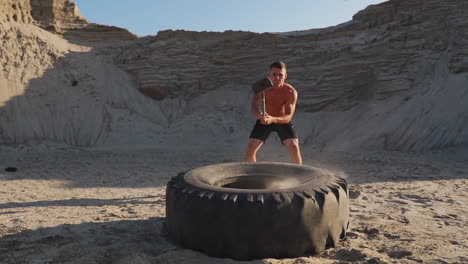 atleta musculoso hombre fuerte golpea un martillo en una rueda enorme en las montañas arenosas en cámara lenta al atardecer. el polvo de las ruedas se eleva.