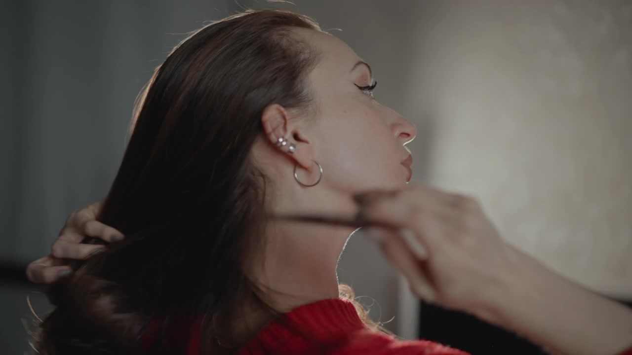 Scena ravvicinata in movimento di una donna con un maglione di maglia rosso che si liscia i lunghi capelli castani con le mani mentre gira la testa verso una luce laterale, creando un'atmosfera di bellezza interna calma con messa a fuoco morbida e texture naturale.