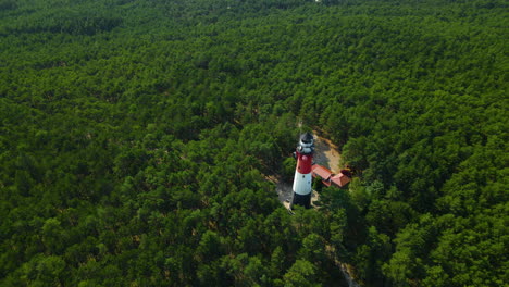 drone flies around red and white lighthouse that stands tall in a lush green forest surrounded by trees