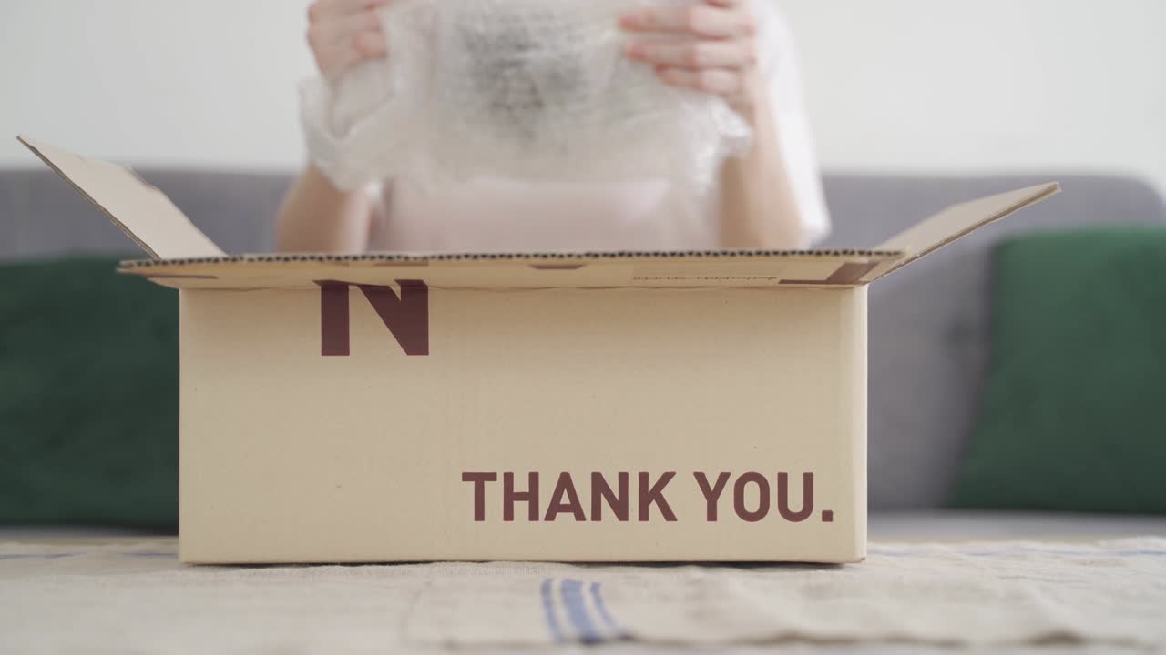 Happy young asian woman opening carton parcel and smiling while sitting on sofa in living room. Cheerful taking out air bubble protection wrap item from cardboard box on couch at home.