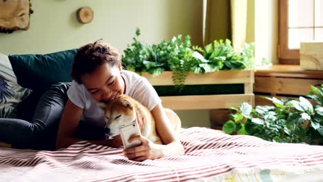 smiling mixed race teenager is taking selfie with puppy then talking to it and showing it photos on smartphone screen lying on bed at home. technology and pets concept.