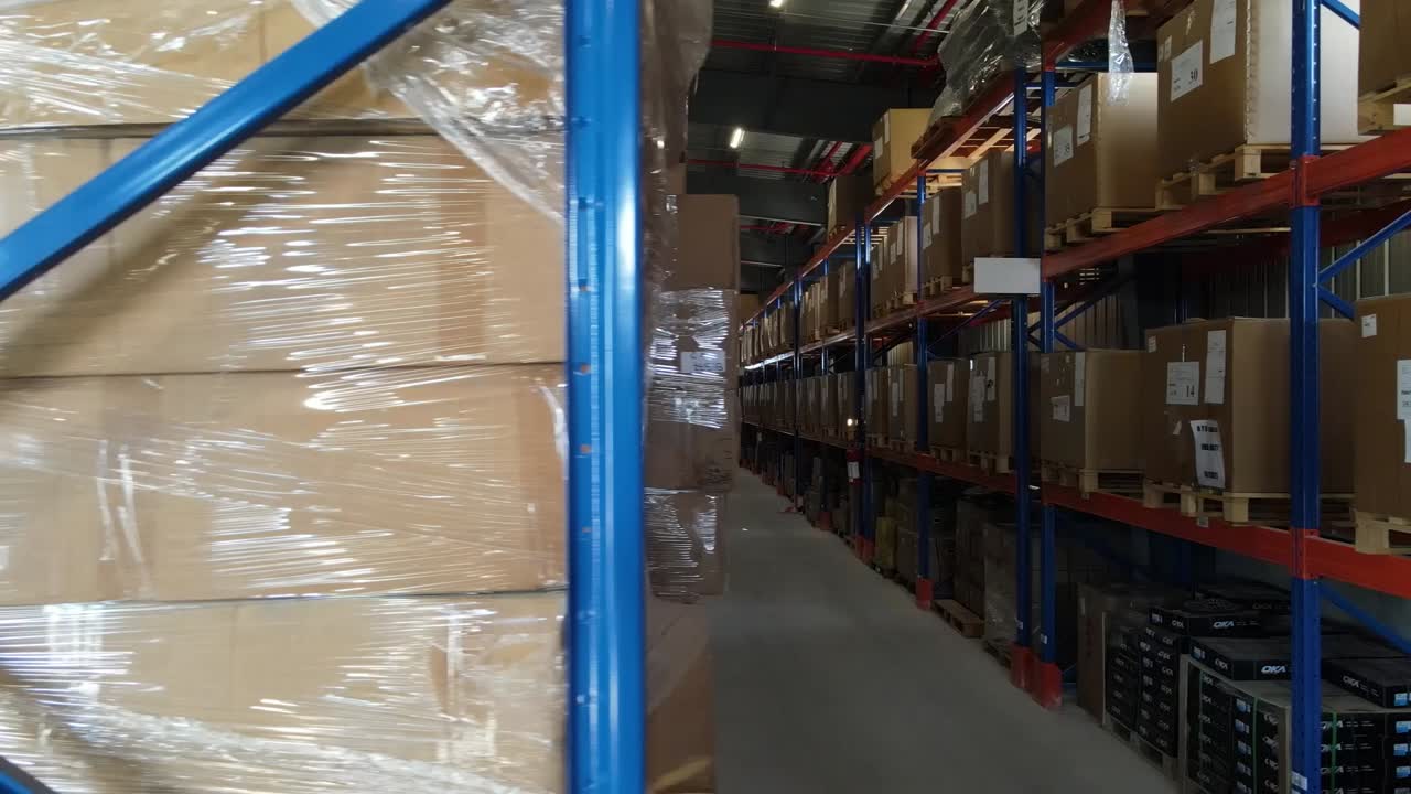 Cardboard boxes wrapped in plastic film are stored on metal shelves in a warehouse with a visible fire extinguisher