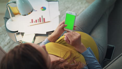 Vista-Superior-De-Una-Mujer-Embarazada-Escribiendo-En-Un-Teléfono-Inteligente-Con-Pantalla-Verde-En-Casa.