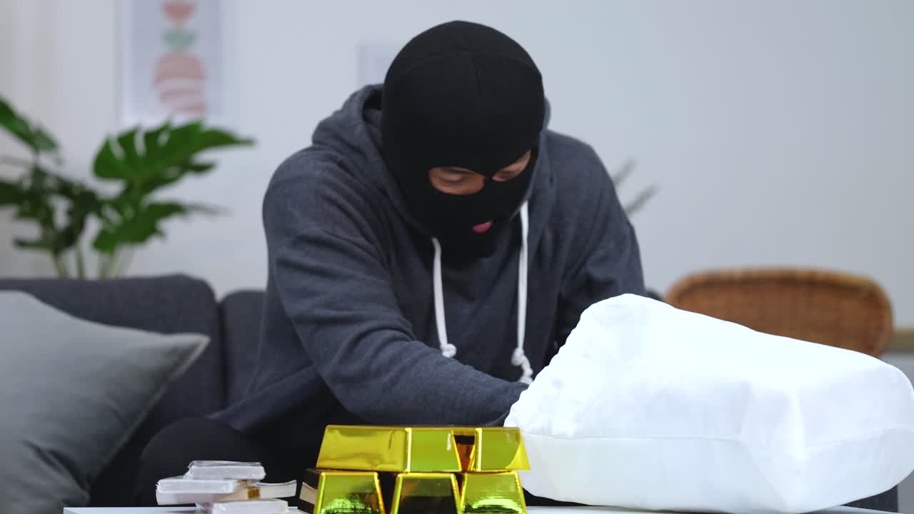 A criminal in a balaclava counts stacks of money beside gold bars and loot
