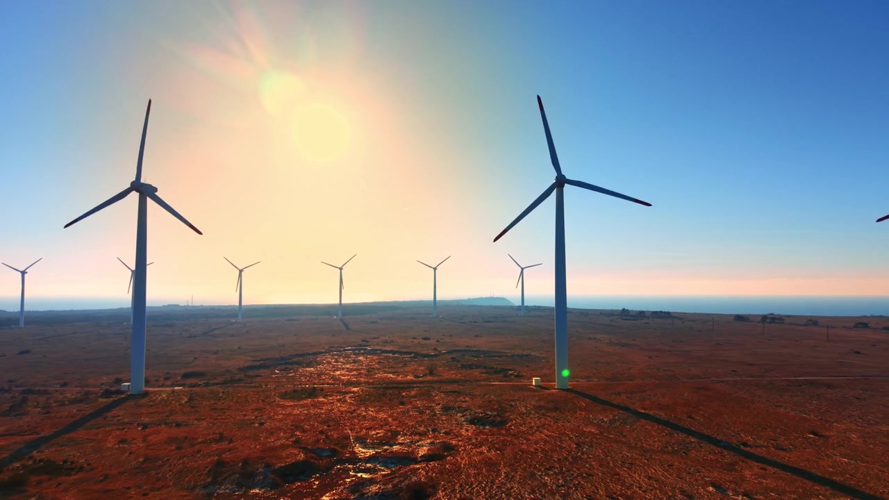Sunrise light over wind farm on red coastal plain. Turbines framing sunburst, long shadows across russet earth, sea glimmering at horizon. Aerial view.