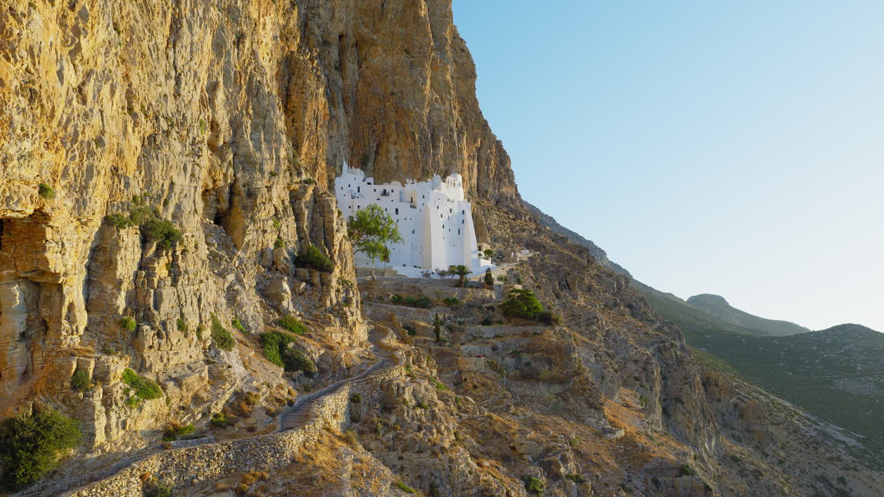 Cinematic drone shot approaching Panagia Hozoviotissa Monastery, gliding along dramatic cliffs bathed in warm morning light