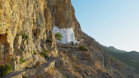 cinematic drone shot approaching panagia hozoviotissa monastery, gliding along dramatic cliffs bathed in warm morning light