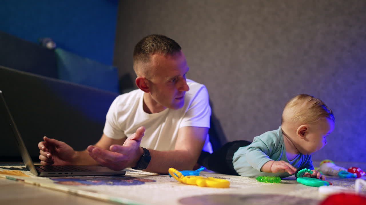 Caucasian father and infant son lying on the floor. Dad works on laptop and baby boy starts to cry.