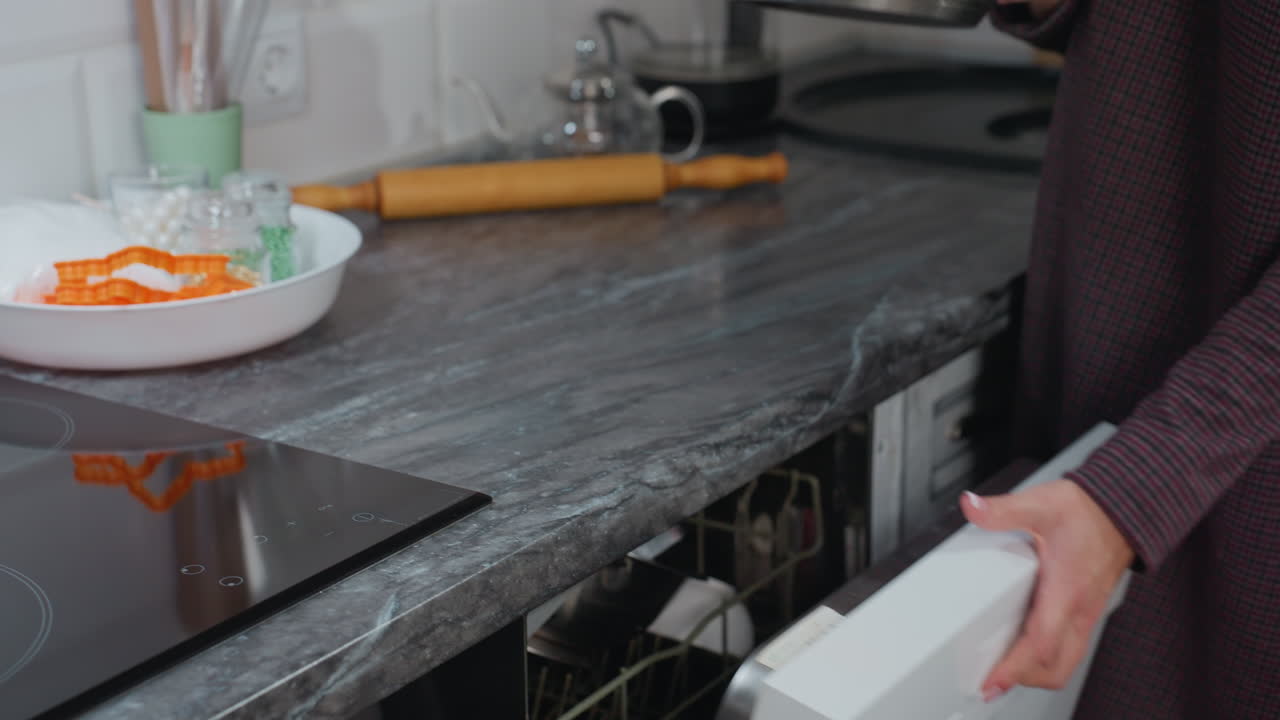 Seorang wanita mengeluarkan panci dari lemari dapur dan meletakkannya di atas kompor listrik modern yang ramping dengan fitur tampilan digital, barang-barang di atas meja, mesin pencuci piring terbuka, interior bergaya, dan suasana rumah tangga yang nyaman