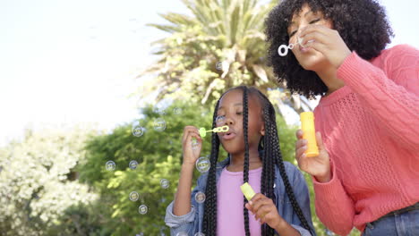 happy african american mother with daughter blowing bubbles in garden at home, slow motion