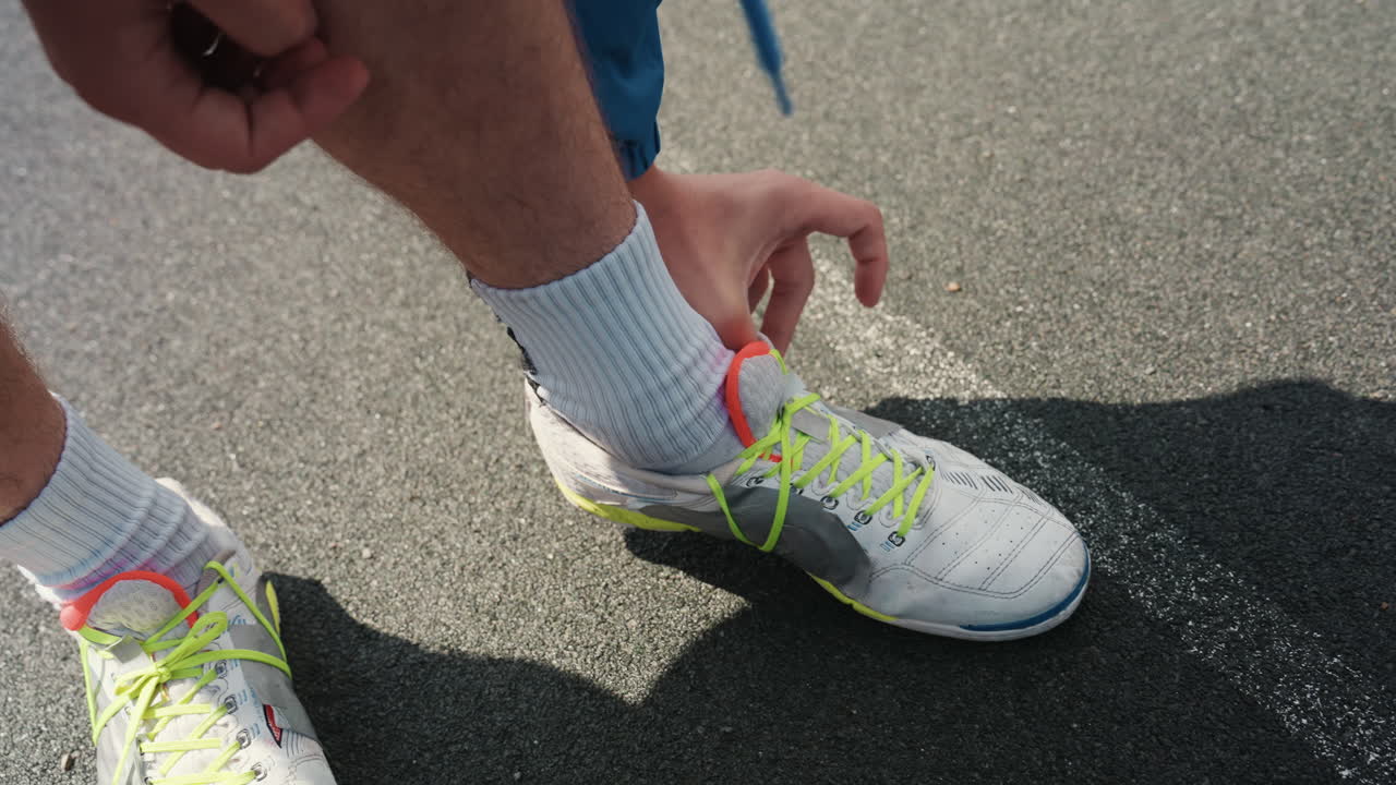 Un giocatore di football americano bianco che si allaccia le scarpe da ginnastica sull'asfalto bagnato, lacci neon e calzini bianchi, mani in primo piano che allacciano le scarpe con polvere di gesso rovesciata e pozzanghere in ombra, rituale pre-partita intenso e pieno di grinta