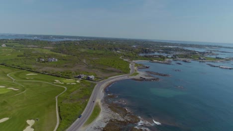 Aerial-of-a-road-next-to-the-ocean-and-a-golf-coast-with-waves-crashing-on-a-rocky-shore-on-a-summer-day