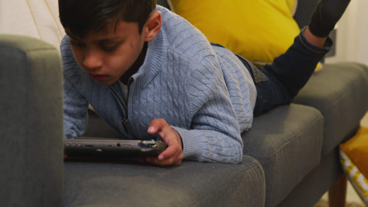 Young Boy Lying On Sofa At Home Playing Game Or Streaming Onto Handheld Gaming Device 4