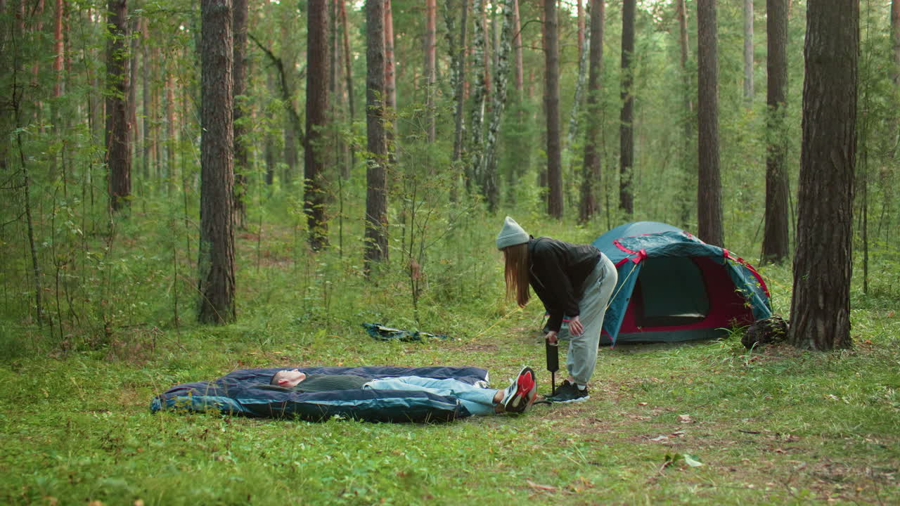 Man lays on tent bag relaxing while woman slightly bent pumps air into inflatable bag with another tent visible behind them in outdoor forest setting surrounded by soft sunlight