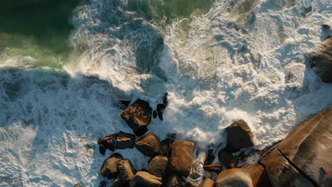 aerial of a guy sitting on a rock watching the waves breaking during sunset in llandudno, cape town, south africa