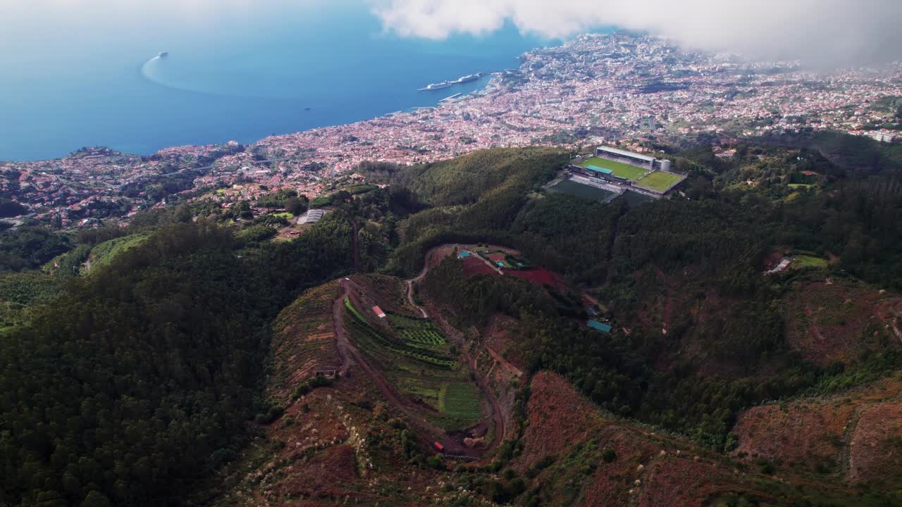 Filmato aereo di Funchal, sull’isola di Madeira, che mostra le verdi colline, il moderno stadio e la costa lungo il blu dell’Oceano Atlantico sotto un cielo nuvoloso, catturando la bellezza scenica del paesaggio dell’isola portoghese.