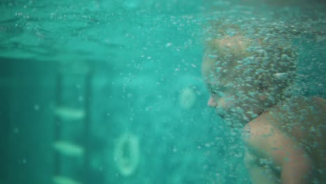 cute blonde toddler is jumping under the water in the swimming pool and swimming there until his mother is lifting him from the water. an underwater shot