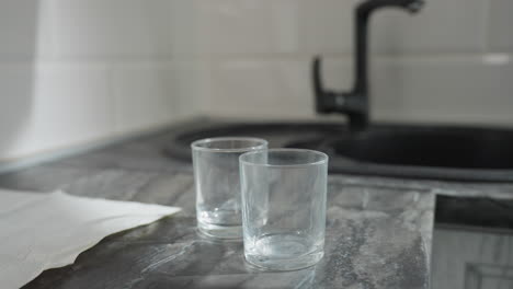 close up of hand gently dropping transparent glass cups on countertop with partial view of tissue paper beside sink in bright modern kitchen interior filled with natural sunlight