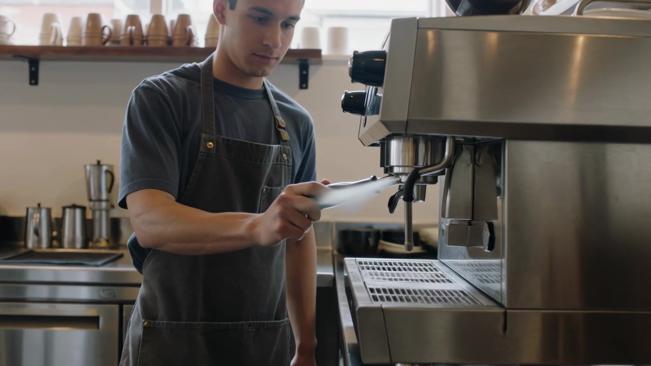 Barista uomo con grembiule scuro che attiva il vapore e allinea il portafiltro al bar, preparando il caffè, con spazio per il testo.