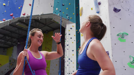 Dos-Mujeres-Caucásicas-Hablando-Y-Chocando-Las-Manos-En-El-Muro-De-Escalada-Interior.