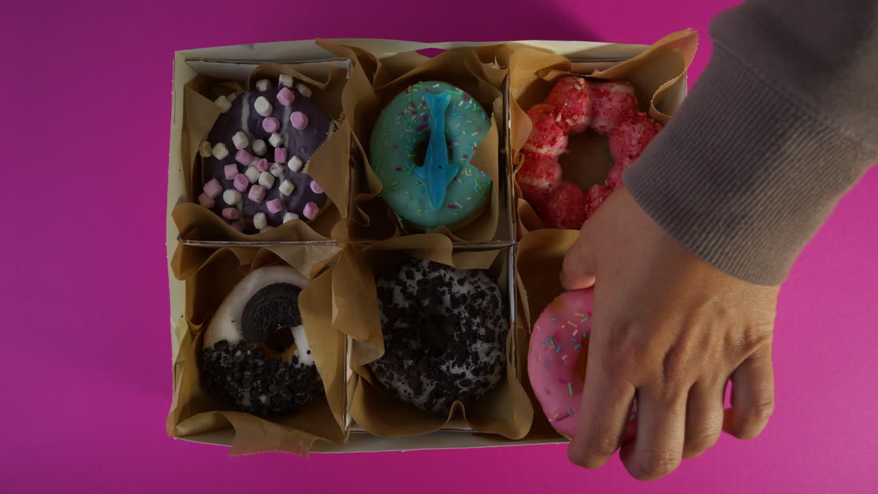 A hand with a gray sleeve places a pink frosted donut with rainbow sprinkles into the final slot of a six-count delivery box, completing a colorful assortment against a bright pink background.